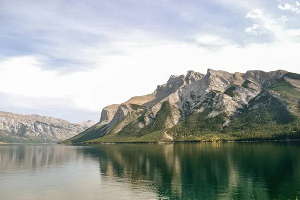 Sakin bir alp gölü yumuşak mavi gökyüzünün altındaki Rocky Dağları 'nın keskin ve engebeli tepelerini yansıtır. Sakin sular kayalıkların karmaşık dokusunu yansıtır ve yamaçlardan aşağı çağlayan yemyeşil ormanları..