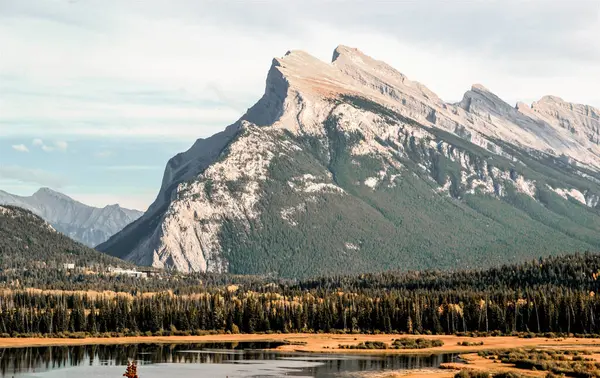 Görkemli Rocky Dağı zirveleri sık yeşil ormanlar ve sakin yansıtıcı göl üzerinde keskin bir şekilde yükselir. Engebeli kaya oluşumları ve canlı yeşillik katmanları sonbaharda nefes kesici bir alp alanı oluşturur..