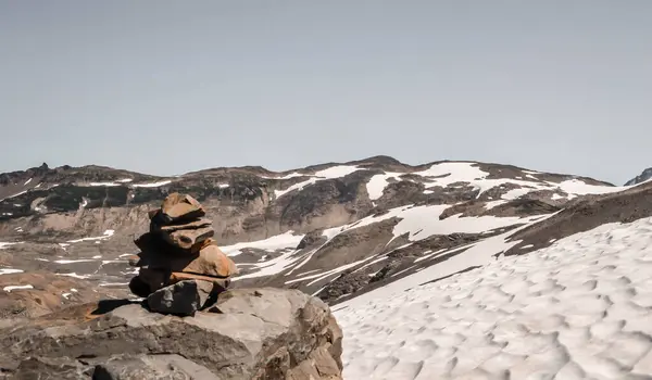 Bir taş cairn, engebeli alp arazisi ve buzullu buz parçalarıyla çevrili, karla kaplı bir dağın tepesinde duruyor. Bu sakin ve minimalist manzara yüksek irtifa keşiflerinin özünü yakalar..