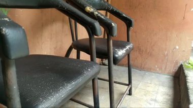 İki siyah sandalye, eski püskü ve kırık bir sandalye. Arka planda evin duvarı var.