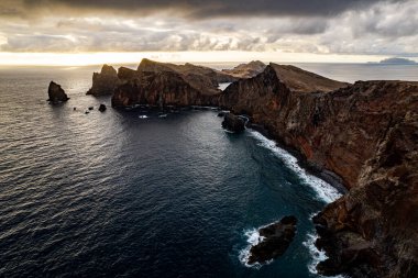 Madeira adasındaki Ponta de Sao Lourenco adlı engebeli bir sahil yarımadasının okyanusla buluşmasının manzarası bulutlu bir gökyüzü altında doğal kaya oluşumları ve dinamik dalgalar sergiliyor. Evcilleştirilmemiş doğanın ve deniz güzelliğinin özünü yakalar.