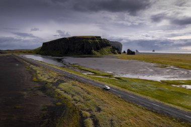 A vehicle travels on a coastal road near blach beach Reynisfjara and lush cliffs, surrounded by dramatic clouds under a moody sky, capturing the beauty of remote, untouched landscapes clipart