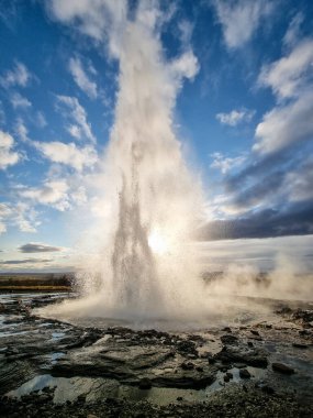İzlanda 'daki Altın Daire' de Geysir 'de canlı bir mavi gökyüzünün altında su ve buharla patlayan bir gayzerin görkemli görüntüsü. Jeotermal faaliyetlerin gücünü ve ihtişamını gözler önüne seren çok güzel bir doğa olayı.