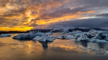 A mesmerizing view of icebergs drifting peacefully in a Jokulsarlon Glacial Lagoon, framed by mountains under a vibrant sunrise sky. The warm sunlight contrasts beautifully with the cold blues of the ice in Iceland clipart