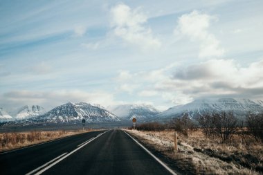 İzlanda 'nın kuzeyindeki geniş bir sıradağ boyunca uzanan ve sakin mavi gökyüzünün altında karla kaplı tepeleri gösteren bir yolun çarpıcı görüntüsü.