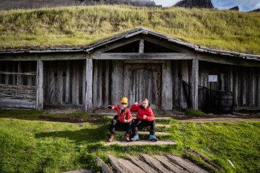 Two individuals wear red jackets sit on the steps of a rustic wooden house in Viking Village in breathtaking Stokksnes in Iceland surrounded by grassy terrain, reflecting a sense of exploration clipart