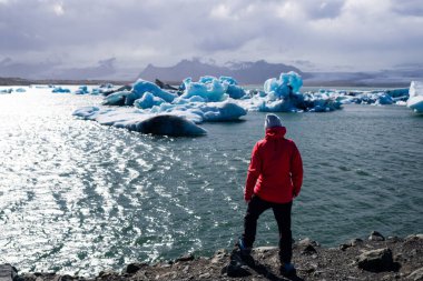 Kırmızı ceket giyen bir kişi, İzlanda 'da Jokulsarlon gölünde yüzen buzdağlarıyla çevrili sakin bir su kütlesine bakar. Bulutlu bir gökyüzü altında, keşif temalarını taşır..