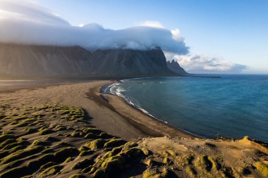 İzlanda 'da siyah kumlu bir plaj, yemyeşil kumullar, altın gün batımı, yüksek Vestrahorn dağları, Atlantik okyanusu ve dramatik bulut oluşumlarının yer aldığı bir kıyı manzarası.