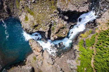 Şelalenin engebeli uçurumlar ve canlı yeşillikler arasında çağlayan Nykurhylsfoss 'un (Sveinsstekksfoss) İzlanda' daki Fossardalur Kamp alanı yakınlarındaki güzelliği yakaladığı nefes kesici bir hava manzarası.