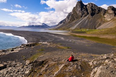 A spectacular aerial drone photograph showcasing couple in red jackets and rugged coastline framed Hvalnes Nature Reserve Beach by dramatic mountain peaks and a vast Atlantic Ocean in Iceland clipart