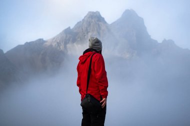 Kırmızı ceketli yalnız bir yolcu, İzlanda 'da nefes kesen Stoksnes' deki sisli Vestrahorn dağlarıyla çevrili kayalık bir arazide duruyor.