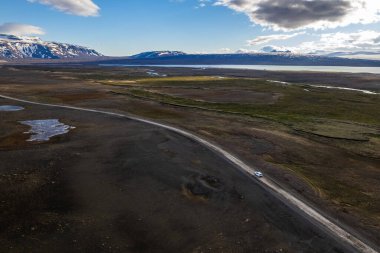 Hava aracı, bulutlu bir gökyüzü altında İzlanda 'nın bu manzaralı bölgesinde seyahat eden tek başına beyaz bir aracın yer aldığı geniş ve çorak bir araziyi kesen uzak bir yola bakıyor.