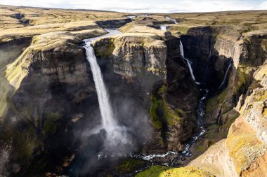 İzlanda 'daki güçlü Haifoss ve Granni şelalelerinin yemyeşil ve kayalık uçurumlarla çevrili derin bir kanyona aktığını gösteren nefes kesici bir hava aracı sahnesi.