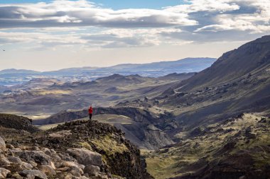 Kırmızı ceketli yalnız bir birey kayalık bir çıkıntıda durup İzlanda 'da geniş ve görkemli bir dağlık çevreye bakıyor. Parçalı bulutlu bir gökyüzünün altında, keşif ruhunu yakalıyor.