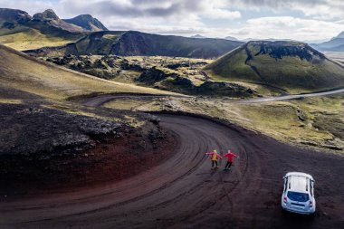 Stunning aerial image showcasing a couple standing over volcanic crater surrounded Landmannalaugar mountains and by hills and wilderness, emphasizing raw beauty and unique geology of volcanic regions clipart