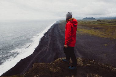 Kırmızı ceketli ve kırmızı şapkalı yalnız gezgin Reynisfjara 'nın derin okyanus manzarası ve dramatik manzaralarla çevrili tepedeki siyah plaj manzarasına hayran.