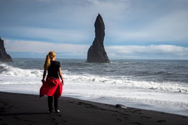 A person walking along a stunning black sandy beach Reynisfjara in Iceland in VIK Myrdal in Golden Circle area with Atlantic Ocean waves crashing and volcanic rock formations in the background clipart