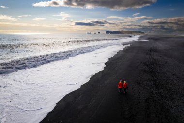 Aerail view couple walking along a stunning black sandy beach Reynisfjara , sunset in Iceland VIK Myrdal in Golden Circle area with Atlantic Ocean waves crashing and volcanic rock formations clipart