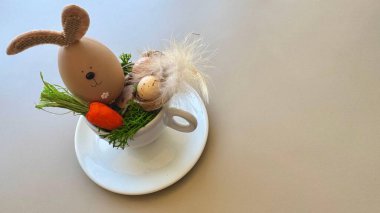 A fun Easter-themed scene featuring a decorative bunny-shaped egg with felt ears nestled in green grass alongside a small carrot, all contained inside a white teacup. clipart