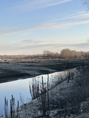 Sakin bir nehir kışın çorak bir arazide, ön planda kuru bitkiler ve uzakta yapraksız ağaçlar arasında süzülür. Yumuşak bir ışık ve berrak bir gökyüzü huzurlu ve düşünceli bir hava yaratır.