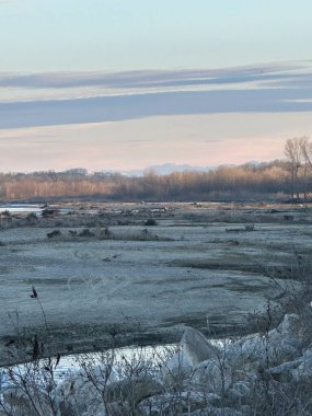 Issız bir kış nehri, yapraksız ağaçlara doğru uzanır ve soğuk, pastel bir gökyüzünün altında soluk dağ sırtlarına. Ön planda, durgunluk ve yalnızlığı çağrıştıran buzlu kayalar ve kuru bitki örtüsü yer alıyor.