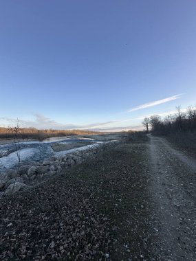 Engebeli bir toprak yol, akan bir kış nehrinin kıyısındaki kayalık bir nehir kıyısını takip ediyor. Etrafı yapraksız bitki örtüsüyle çevrili ve uçsuz bucaksız mavi gökyüzünün altında altın renkli ağaçlarla çevrili.