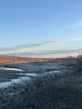 Geniş, çoğunlukla kuru bir nehir yatağı kışın seyrek bitki örtüsü ve yapraksız ağaçlarla dolu bir arazide esiyor. Açık mavi gökyüzü ve yumuşak sabah ışığı bu doğal manzaraya huzur katar.