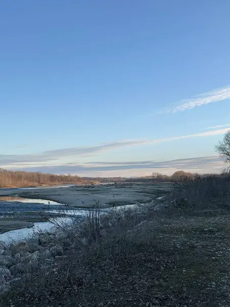 Kuru çimenler, yapraksız ağaçlar ve yumuşak mavi bir gökyüzünün altındaki kayalık kıyılar arasında kıvrılan sığ bir nehrin geniş manzarası. Sakin ve sakin doğal bir sahne.
