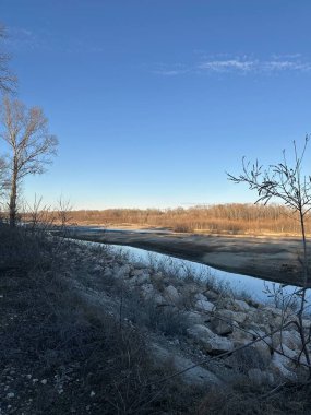 Sakin bir nehir, çıplak ağaçlar ve kuru çalılarla çevrili soğuk bir kış manzarasında esiyor. Berrak mavi gökyüzü durgun suya yansıyarak sakin ve minimalist bir manzara yaratır.