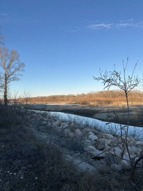 Huzurlu bir kış nehri çorak bir arazide kıvrılır, yapraksız ağaçlar yumuşak altın ışığı yansıtır. Açık mavi gökyüzü ve kuru bitki örtüsü dinginlik ve yalnızlık hissini arttırır.