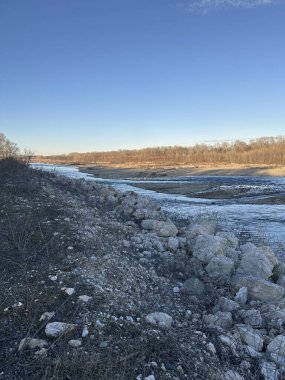Kış bitkileriyle ve yapraksız ağaçlarla çevrili kayalık bir arazide dolambaçlı bir nehir akar. Yumuşak altın ışık, bulutsuz mavi gökyüzünün altında ormanı yıkar.