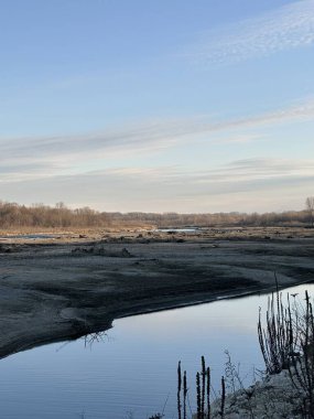 Sakin bir kış nehri, çorak bir arazide süzülürken gökyüzünü yansıtır. Durgun su, kuru bitkiler ve yumuşak ışık huzuru ve mevsimsel durgunluğu çağrıştırıyor.