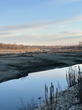 Sakin bir dere, solgun bir kış gökyüzünün altındaki soğuk bir arazide esiyor. Suyun ve kuru bitkilerin yansımaları sakin bir mevsimsel atmosfer yaratır.