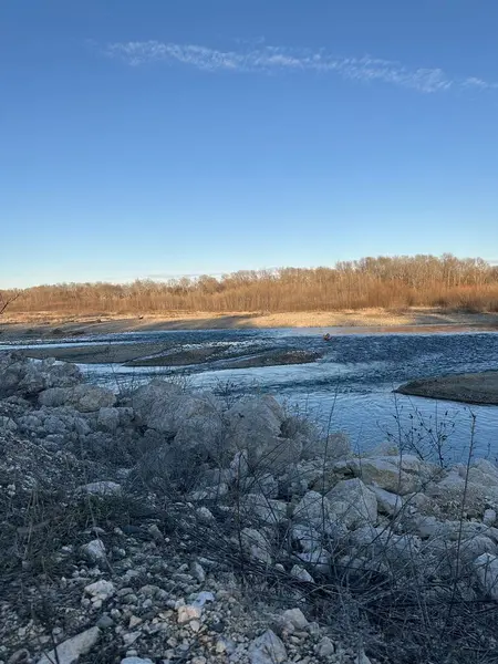 Kış gökyüzünün altında sığ bir nehir kayalık bir kıyı şeridini geçer. Arka plandaki çıplak ağaçlar ve altın ışık ön plandaki su ve taşların serin tonlarıyla tezat oluşturuyor.