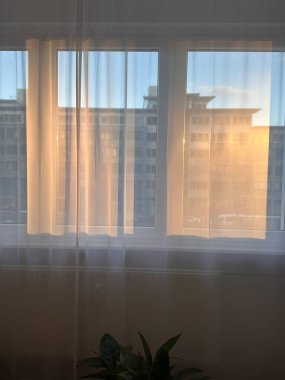 Saf beyaz perdelerden süzülen yumuşak altın güneş ışığı arka planda şehir manzarası olan modern bir pencereye sıcak bir ışık saçıyor. Huzurlu, minimal sabah sahnesi