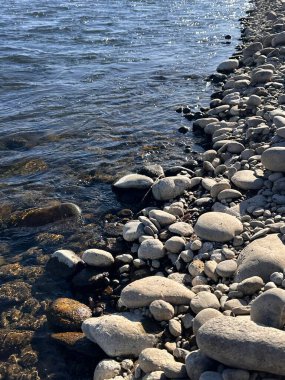 Yumuşak çakıl taşlarının ve taşların doğal güneş ışığı altında dalgalanan suyla buluştuğu kayalık bir kıyı şeridi manzarası. Doğa, sükunet ve manzara temaları için ideal