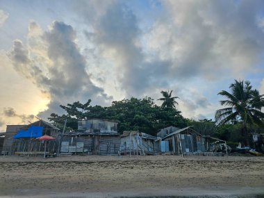 Görüntü, alacakaranlıktaki sahil manzarasını gösteriyor. Sahile yakın kırsal ahşap ve metal evler inşa edilmiş. Bazı yapılar mavi ve kırmızı muşambalarla kaplıdır. Sahilin ıslak kumu ön planda belirir.. 