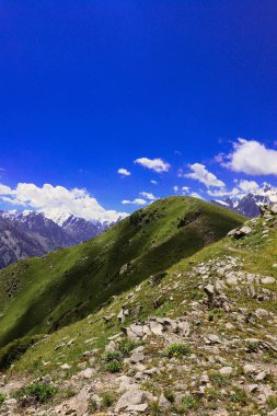 Fotoğrafta, açık mavi gökyüzünün altında dağları görüyoruz, beyaz bulutlarla dağılmış.