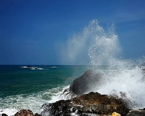 Üzerinde dingin mavi gökyüzü olan engebeli kıyı kayalarına vuran canlı okyanus dalgaları, deniz kıyısının doğal güzelliğini ve dinamik enerjisini sergiliyor..