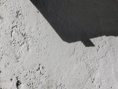Güneş ışığıyla aydınlanan desenli beyaz bir duvar. Keskin, geometrik bir gölge bırakıyor. Işığın ve gölgenin etkileşimi duvarın düz olmayan yüzeyini vurgular.. 