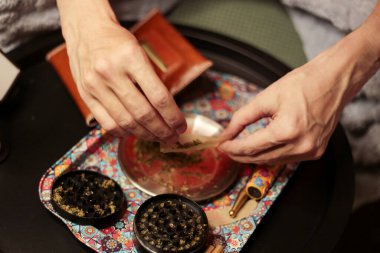 Bir esrarı dikkatlice sarmak için esrarı renkli bir tepsinin üzerinde sarmak. Sahnede bir öğütücü, bir kese ve dingin ve odaklanmış bir sigara piposu var.