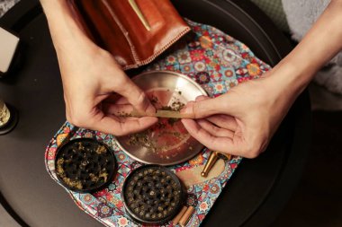 Bir esrarı dikkatlice sarmak için esrarı renkli bir tepsinin üzerinde sarmak. Sahnede bir öğütücü, bir kese ve dingin ve odaklanmış bir sigara piposu var.