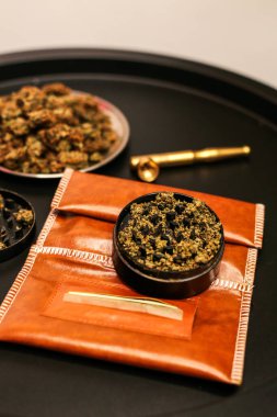 Kahverengi deri bir kesede içi ezilmiş kenevirle dolu bir öğütücüye yakın çekim. Yuvarlanan bir kağıt küçük bir kompartımana yerleştirilir. Tomurcukları ve arka planda altın bir borusu vardır.