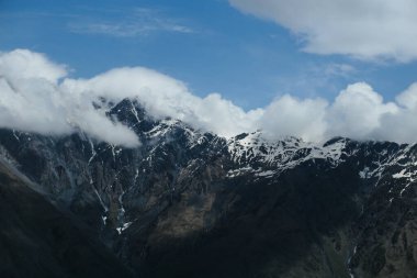 Kafkas Dağları 'nın engebeli ve karla kaplı zirvelerinin yakın görüntüsü kısmen mavi bir gökyüzünün altında kabarık bulutlarla kaplıdır ham ve görkemli bir alp manzarası..