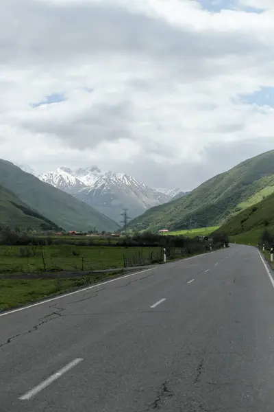 Geniş bir asfalt yol, Kafkasya Dağları 'ndaki bulutlu bir yay gökyüzünün altında, karla kaplı tepeleri olan yemyeşil bir vadide esiyor..
