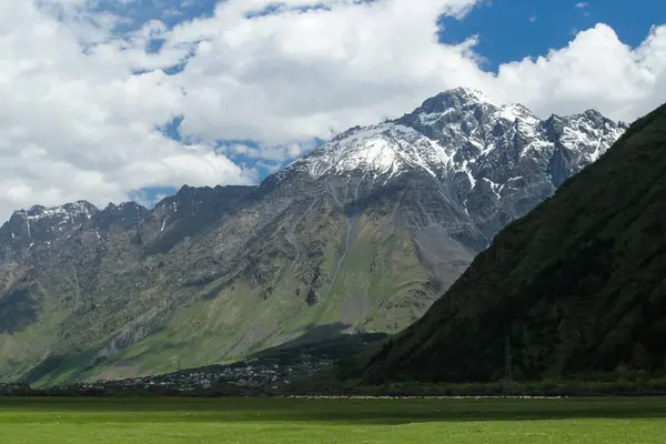 Gürcistan 'ın Kazbegi kentindeki Kafkas Dağları' nın görkemli kar tozlu zirveleri yemyeşil bir vadinin arkasında yükseliyor. Dramatik uçurumlar, aşağıdaki yumuşak otlaklarla çok güzel tezat oluşturuyor..