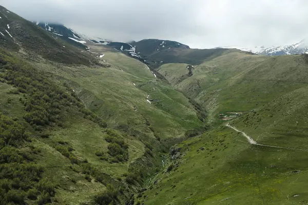 Dağılmış kar parçaları ve dramatik bulutlarla yemyeşil bir dağ vadisinin panoramik manzarası. Issızlığı ve macerayı çağrıştıran dar bir patika, çölde yol açar..