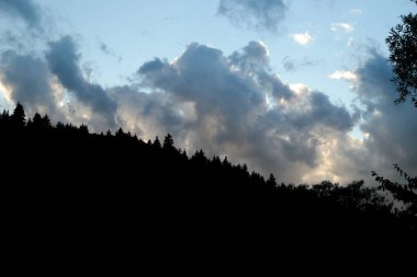Dark forest silhouettes contrast with a dramatic sky filled with thick clouds illuminated by the setting sun, creating a moody mountain evening atmosphere.
