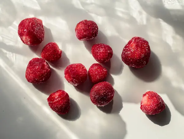 Close-up of frozen strawberries on a white surface with soft natural shadows, highlighting their red texture and frosty details.