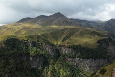 A panoramic view of a mountain valley with steep rocky slopes, green meadows, and cloudy sky above.